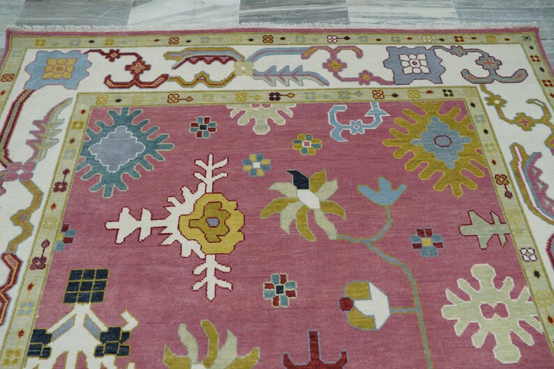 May include: A detailed shot of a decorative rug with a pink central field. The rug features a complex floral pattern in white, yellow, blue, and red, with a cream-colored border. The design is symmetrical and intricate.