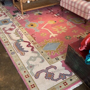 May include: A large, hand-knotted rug with a pink field and a cream-colored border. The rug features floral and geometric patterns in shades of blue, yellow, and green. The rug is displayed in a room with furniture.