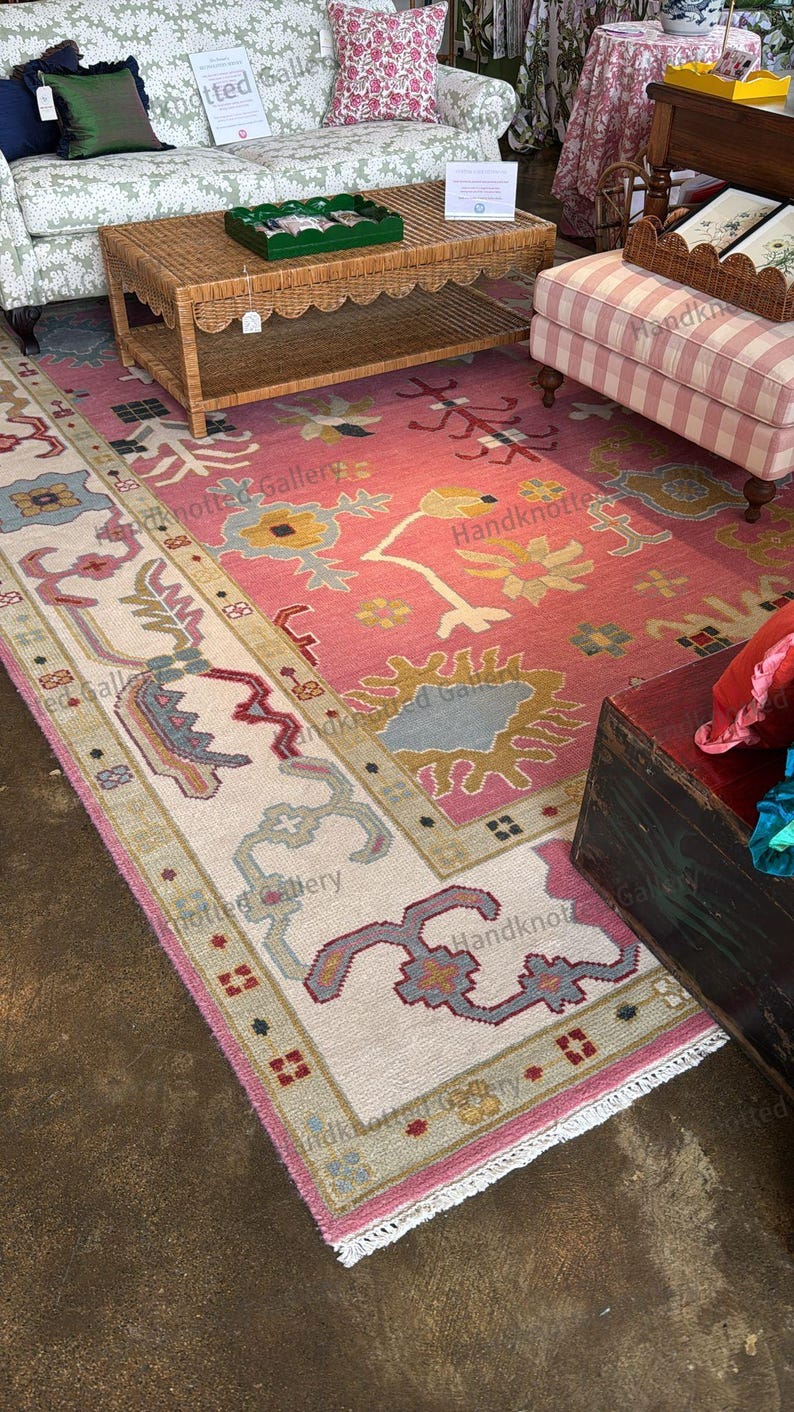 May include: A large, hand-knotted rug with a pink field and a cream-colored border. The rug features floral and geometric patterns in shades of blue, yellow, and green. The rug is displayed in a room with furniture.
