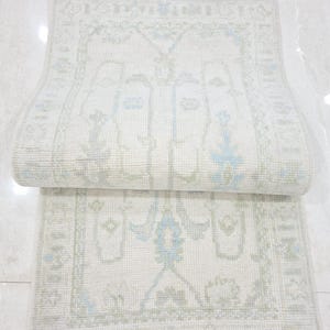 Puede incluir: Una alfombra blanca rota plegada con un patrón detallado en azul claro y verde. La alfombra tiene forma rectangular con flecos en los bordes. El diseño presenta elementos geométricos y florales.