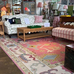May include: A showroom featuring a variety of home decor items. A pink and cream-colored rug with a floral design is on the floor. A wicker coffee table and a floral patterned sofa are in the center. Several lamps with patterned shades are visible.