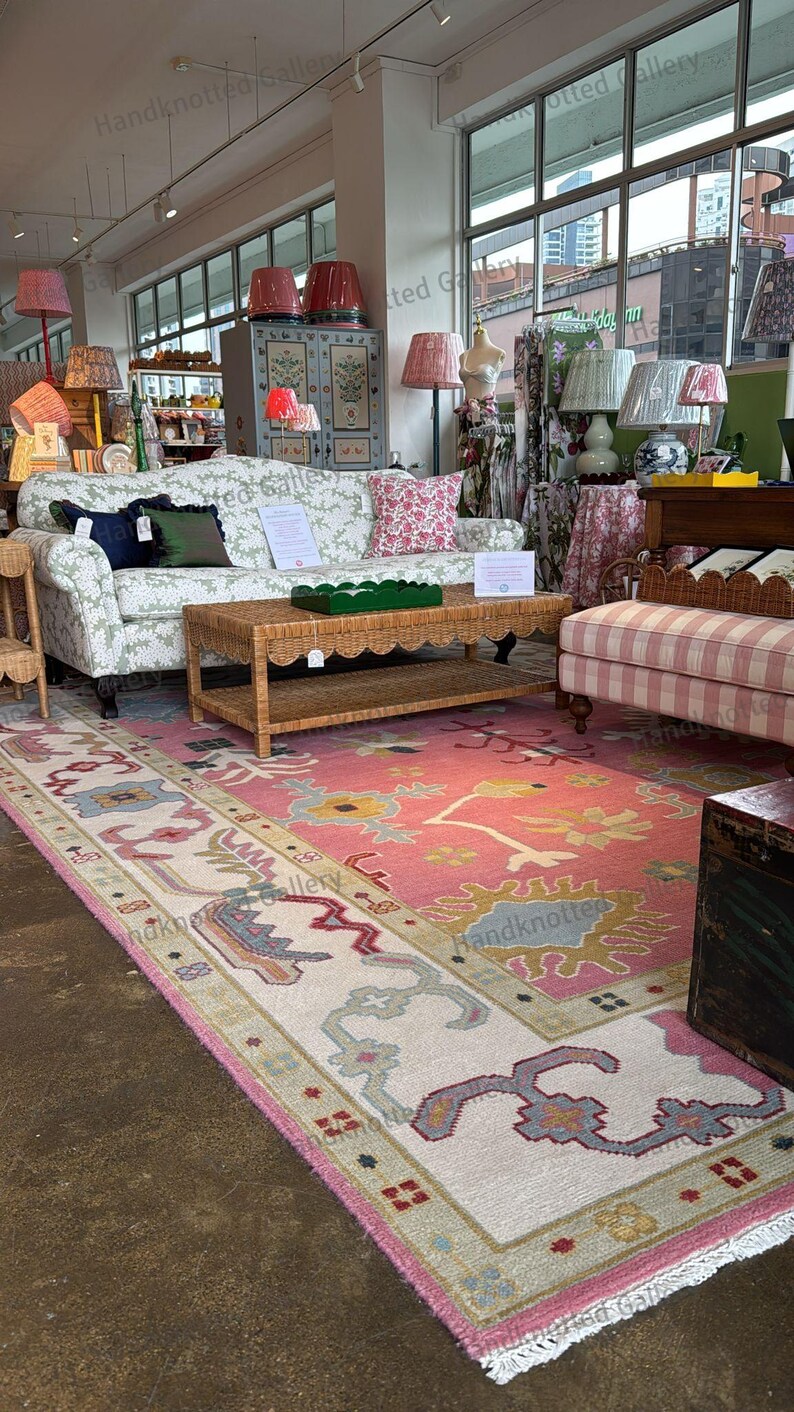 May include: A showroom featuring a variety of home decor items. A pink and cream-colored rug with a floral design is on the floor. A wicker coffee table and a floral patterned sofa are in the center. Several lamps with patterned shades are visible.