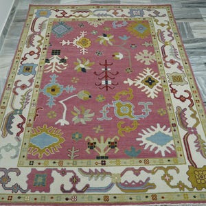 May include: A rectangular rug with a rose-colored center and a cream-colored border. The rug features a repeating pattern of stylized floral and geometric designs in shades of blue, yellow, and green. The rug is placed on a gray tiled floor.