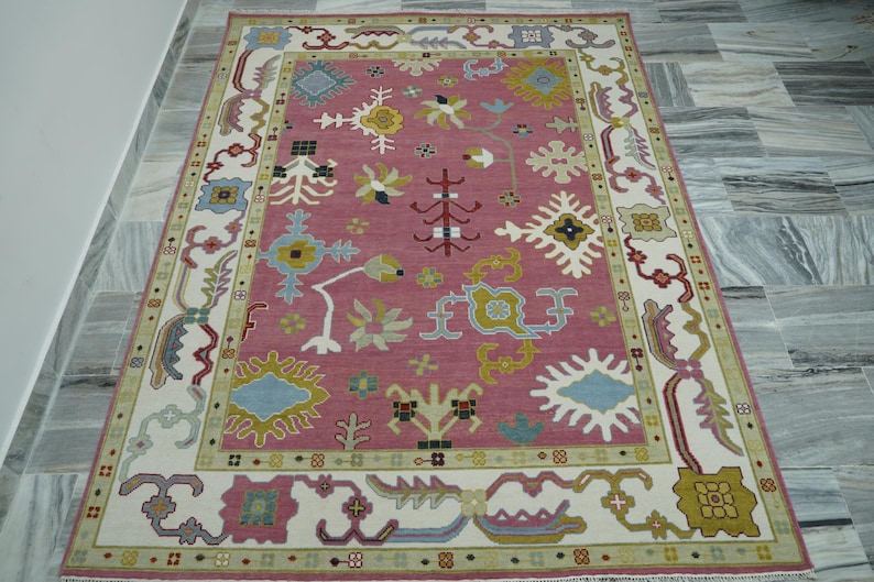 May include: A rectangular rug with a rose-colored center and a cream-colored border. The rug features a repeating pattern of stylized floral and geometric designs in shades of blue, yellow, and green. The rug is placed on a gray tiled floor.