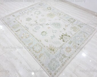 May include: A rectangular, off-white rug with a floral pattern and a light blue border. The rug features a central design of various floral and botanical elements in shades of green and beige. The rug is placed on a white tiled floor.
