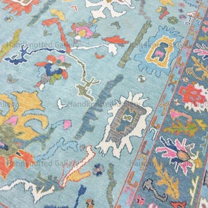May include: A hand-knotted rug with a light blue background and a complex pattern of colorful shapes. The design includes orange, yellow, pink, and blue elements, with a detailed border. The rug's texture is visible.