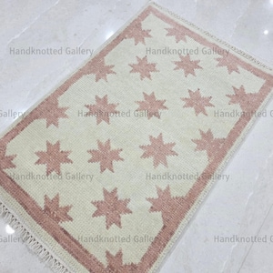 May include: A rectangular rug with a cream-colored background and a repeating pattern of pink stars. The rug has a pink border and fringe along the edges. The words "Handknotted Gallery" are printed on the rug.