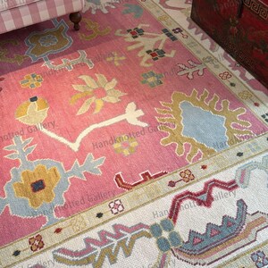 May include: A pink and ivory hand-knotted rug with a floral pattern. The rug features a central field of pink with stylized floral motifs in yellow, blue, and white. The border is ivory with complementary designs. The rug is a decorative floor covering.