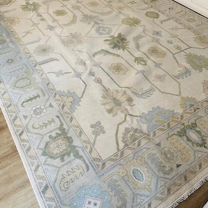 May include: A large, rectangular area rug with a floral pattern in shades of cream, blue, and green. The rug features a detailed border and a central design of stylized flowers and vines. The rug is placed on a hardwood floor.