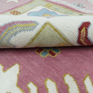 May include: A rolled-up, patterned rug with a pink and white color scheme. The rug features a repeating floral design with accents of yellow, blue, and red. The rug's texture is visible, suggesting a woven construction. The overall aesthetic is traditional.