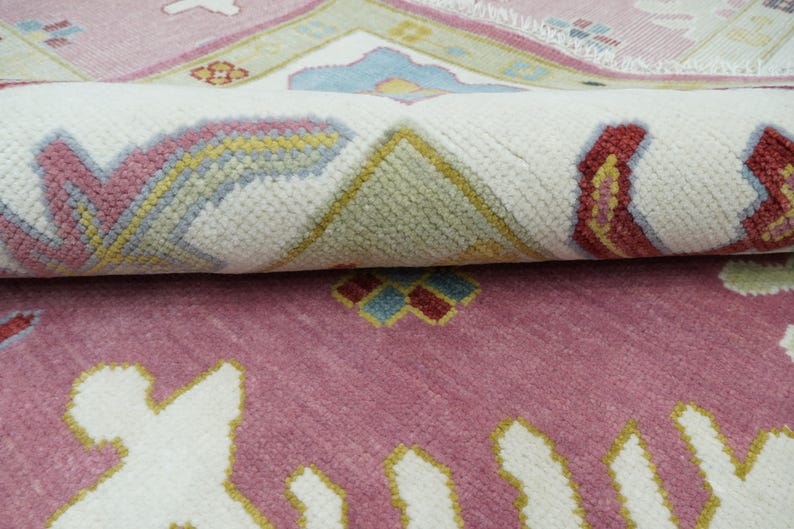 May include: A rolled-up, patterned rug with a pink and white color scheme. The rug features a repeating floral design with accents of yellow, blue, and red. The rug's texture is visible, suggesting a woven construction. The overall aesthetic is traditional.