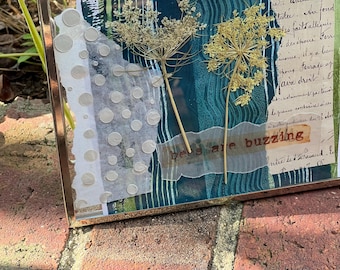 May include: A rectangular metal frame containing a mixed-media collage with two dried, light-colored wildflower stems. The artwork features layers of teal and blue patterned paper, white translucent paper with circular cutouts, and text.