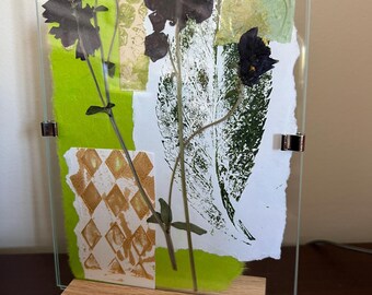 May include: A rectangular glass frame held by a light wood base, displaying a collage of pressed dark purple flowers, green paper, and white paper with green leaf prints and gold diamond patterns, secured by three silver metal clips.