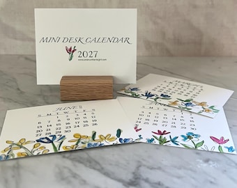 May include: A 2027 mini desk calendar set with a wooden block stand. The cover card features the text MINI DESK CALENDAR 2027 and a small floral illustration. Other cards display monthly grids for June and March with colorful floral borders.