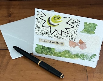 May include: A white folded greeting card with a collage design featuring a stylized lime green and black flower, green textured paper accents, and the text Live Love Grow printed on a small strip. A black fountain pen rests in front of the card.