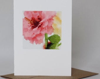 May include: A white greeting card featuring a watercolor painting of a pink flower with green leaves. The card is propped up on a brown envelope. The artist's name and studio name are printed at the bottom of the card.