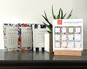 May include: A 2027 desk calendar set featuring a wooden stand and multiple loose-leaf pages. The front page displays a grid of twelve months with colorful floral and geometric borders. Additional pages show November, April, and January with botanical designs.