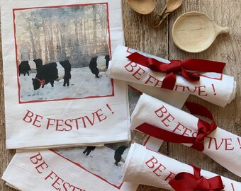 May include: Four white tea towels with red text that reads "Be Festive!" and a watercolor illustration of black and white cows in a snowy forest. The towels are tied with red ribbon.