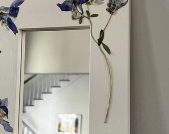May include: A rectangular wall mirror with a wide, off-white wooden frame. The frame is decorated with several pressed, dried blue columbine flowers with green stems and leaves attached to the surface of the wood, extending onto the mirror glass.