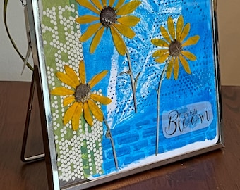 May include: A square metal frame containing a mixed media art piece with three pressed yellow flowers against a bright blue background. The artwork features a green patterned strip on the left and the text "In full Bloom" in the bottom right corner.