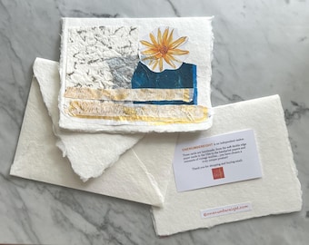 May include: A handmade greeting card with a deckle edge, featuring a collage of textured white, blue, and gold paper with a yellow flower illustration. It includes a matching envelope and a small white card with the text ONENUMBEREIGHT is an independent maker.