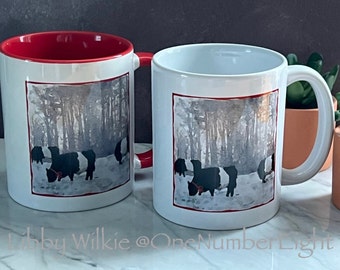 May include: Two white ceramic mugs with red interior rims. Each mug features a watercolor illustration of black and white cows in a snowy forest. The text "Libby Wilkie @ One Number Eight" is printed on the bottom of the image.