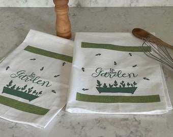 May include: Two white kitchen towels with green text and images. The text on the towels reads "In the Garden".