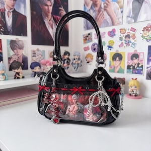 May include: Black patent leather handbag with a clear front panel displaying a graphic. The bag features red bows, a decorative charm, and silver chain accents. The handles are black and the bag is sitting on a white surface.