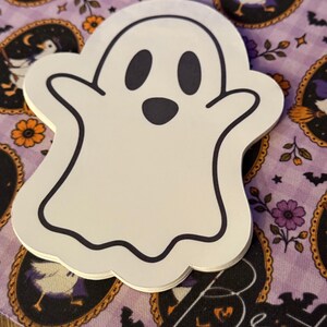 Op de afbeelding: Een stapel gestanste stickers in de vorm van een witte geest met een zwarte omlijning, twee ovale ogen en een kleine ronde mond. De geest heeft afgeronde armen die naar buiten zijn gericht en een golvende onderrand met een simpel ontwerp.