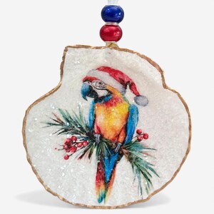 May include: A Christmas ornament featuring a colorful parrot wearing a Santa hat, perched on a branch with red berries and green leaves. The ornament is in the shape of a shell with a gold trim and a beaded string for hanging.
