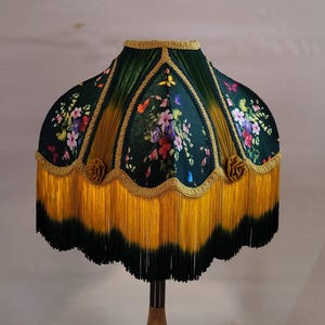 Può includere: Paralume decorativo con un motivo floreale verde scuro. Presenta finiture dorate e una lunga frangia gialla con bordo verde scuro. Il paralume è un accessorio decorativo per la casa. Il motivo floreale comprende fiori rosa, viola e gialli.