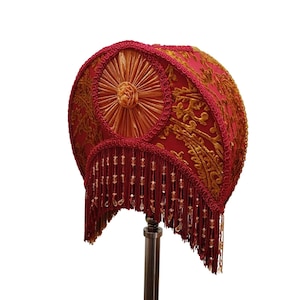 May include: A red lampshade with gold embroidery and a fringe. The lampshade has a circular design with a central rosette and detailed patterns. This decorative lampshade would add a touch of vintage elegance to any room.