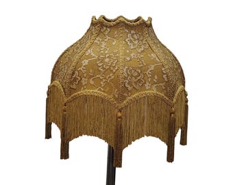May include: A vintage-style lampshade with a scalloped edge and a floral lace pattern in gold and cream. The lampshade features a fringe trim with tassels, adding a decorative element. The lampshade is designed to fit on a lamp base.