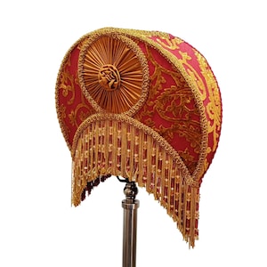 May include: An ornate lampshade in red and gold, featuring detailed floral embroidery and beaded fringe. The lampshade has a circular design with a central rosette and is trimmed with gold braid. The style is vintage and decorative.
