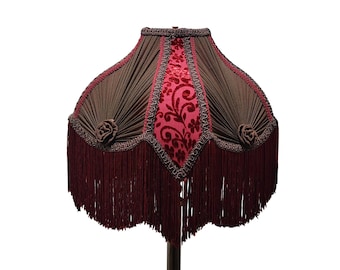 Puede incluir: Pantalla de lámpara decorativa con tela plisada marrón oscuro y un panel central de terciopelo rojo con un diseño floral. La pantalla está adornada con flecos rojo oscuro y rosetas decorativas.