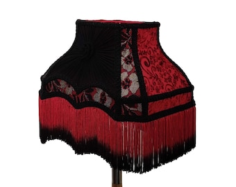 May include: A decorative lampshade with a black top and a red patterned fabric. The lampshade has a fringe of red tassels along the bottom edge. The lampshade is designed to fit on a lamp base.