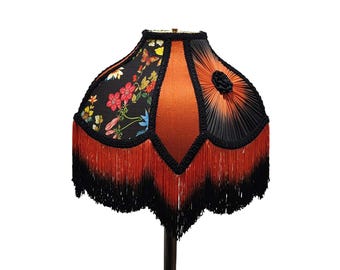 May include: A decorative lampshade with a black, orange, and red color scheme. The lampshade features a floral pattern on a black background, orange center, and black fringe. The lampshade is a unique home decor item.