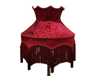 May include: A red lampshade with a floral pattern and fringe trim. The lampshade has a scalloped top and a flared shape. The fringe hangs from the bottom edge of the shade. The lampshade is a decorative lighting element.