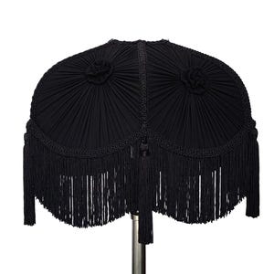 Puede incluir: Una pantalla de lámpara negra con un diseño plisado y flecos. La pantalla tiene forma redondeada con dos acentos florales decorativos y una borla. La franja cuelga del borde inferior.
