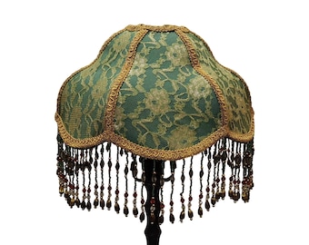 Puede incluir: Pantalla de lámpara verde con un patrón de encaje floral y ribete dorado. La pantalla tiene un borde festoneado y está adornada con flecos de cuentas en tonos marrón, rojo y dorado. Lámpara decorativa.