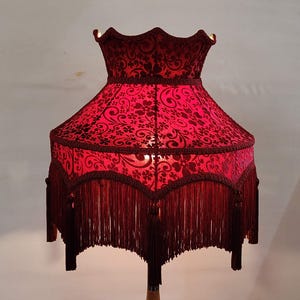 Puede incluir: Una pantalla de lámpara roja con un estampado floral y flecos. La pantalla tiene una parte superior festoneada y una forma acampanada. La franja cuelga del borde inferior de la pantalla. La lámpara es de estilo vintage.