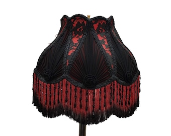 Puede incluir: Una pantalla de lámpara decorativa con una combinación de colores negro y rojo. La pantalla presenta un borde festoneado, flecos negros y detalles de encaje negro. La pantalla tiene un diseño plisado y está adornada con pequeñas rosetas decorativas.