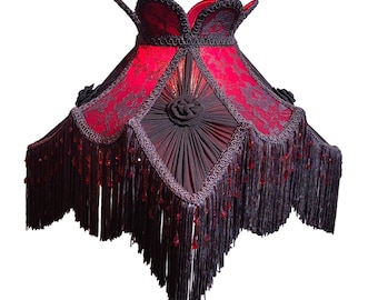May include: A decorative lampshade with a red and black color scheme. The lampshade features a red lace design, black fringe, and decorative accents. The shade has a unique shape and is designed to be placed on a lamp base.