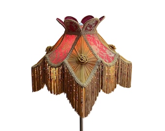 Op de afbeelding: Een vintage lampenkap met een gelaagd ontwerp. De kap heeft een rood en goud bloemmotief, gouden franjes en decoratieve rozetten. Een uniek woondecoratie-item.
