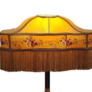 May include: A vintage-style lampshade with a yellow fabric top and a floral patterned band. The band features red and pink flowers. The lampshade has a brown fringe and trim. The lamp is on a black and brown pole.
