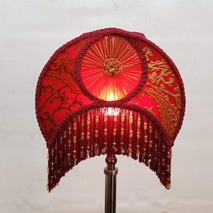 Puede incluir: Una pantalla de lámpara roja con detalles bordados en dorado y una borla de cuentas. La pantalla tiene forma semicircular con un diseño circular central. La tela tiene un estampado de cachemira. La lámpara está sobre un pie de metal plateado.
