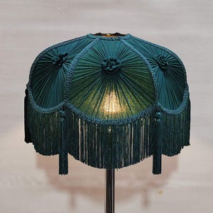 Op de afbeelding: Een tealkleurige lampenkap met een geplooid ontwerp en franjes. De kap heeft decoratieve rozetten en een geschulpte rand. De lamp heeft een verchroomde metalen voet en het licht schijnt door de kap.