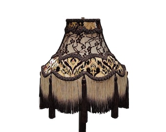 Op de afbeelding: Een decoratieve lampenkap met een donkerbruine kanten overlay en een patroonstof. De kap heeft een geschulpte rand met franjes en kwastjes, wat een vintage esthetiek creëert. Het kleurenpalet omvat bruin, beige en goud.