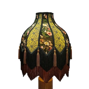 Könnte beinhalten: Ein glockenförmiger Lampenschirm aus leuchtend grünem Stoff mit einem Blumenmuster aus weißen und orangefarbenen Blüten sowie grünen Blättern. Der Schirm ist mit schwarzer geflochtener Borte verziert und hat lange, geschichtete Fransen.