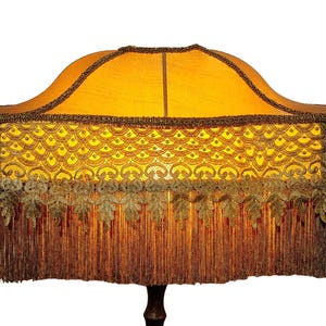 Puede incluir: Pantalla de lámpara de estilo vintage con tela amarillo dorado. Presenta intrincados adornos decorativos, incluyendo un patrón festoneado y detalles en forma de hojas. Largas y marrones flecos cuelgan de la parte inferior, añadiendo un toque clásico.